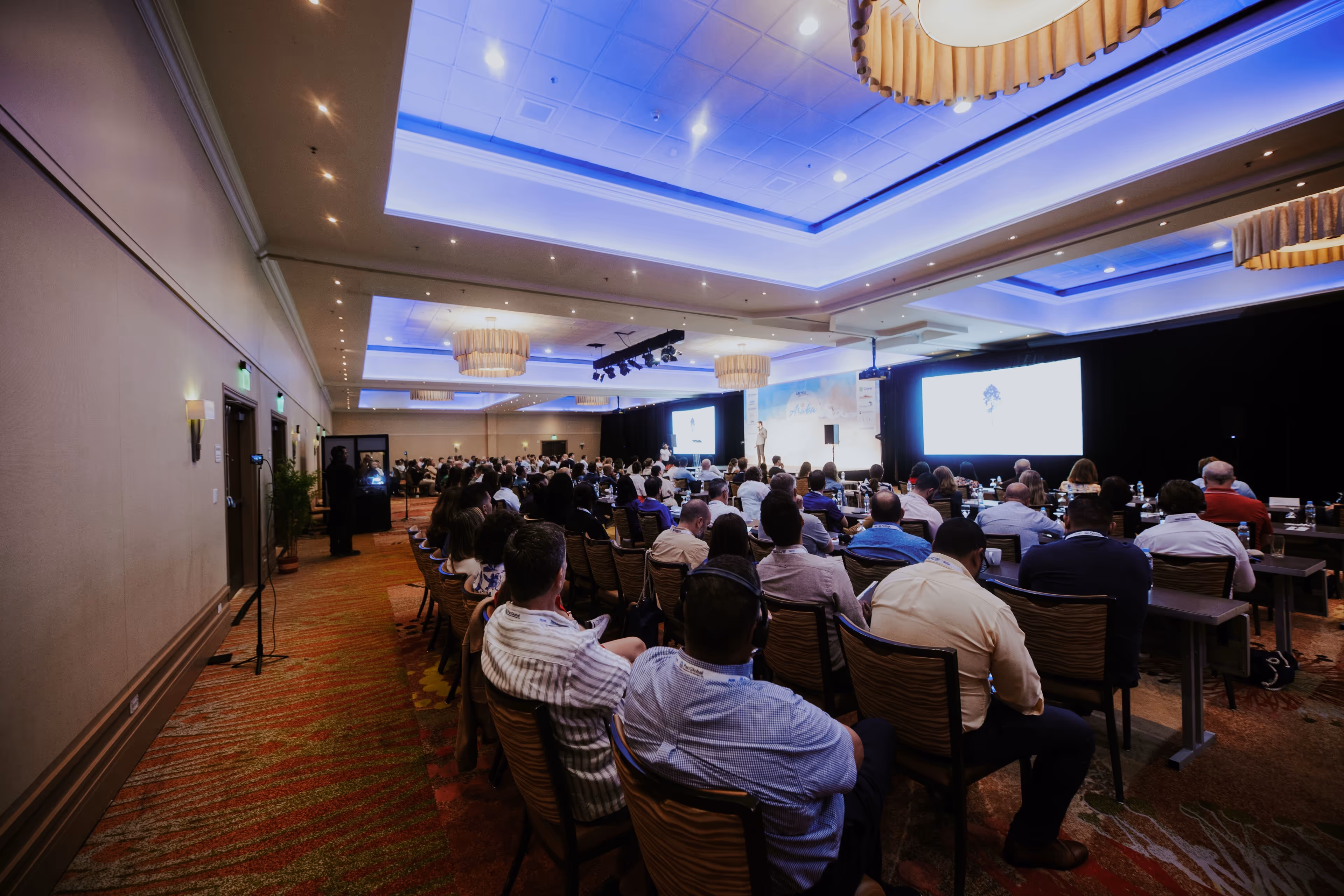 A wide shot from the back of a large conference room showing an audience seated at tables and listening to a speaker on a stage with two large projection screens.