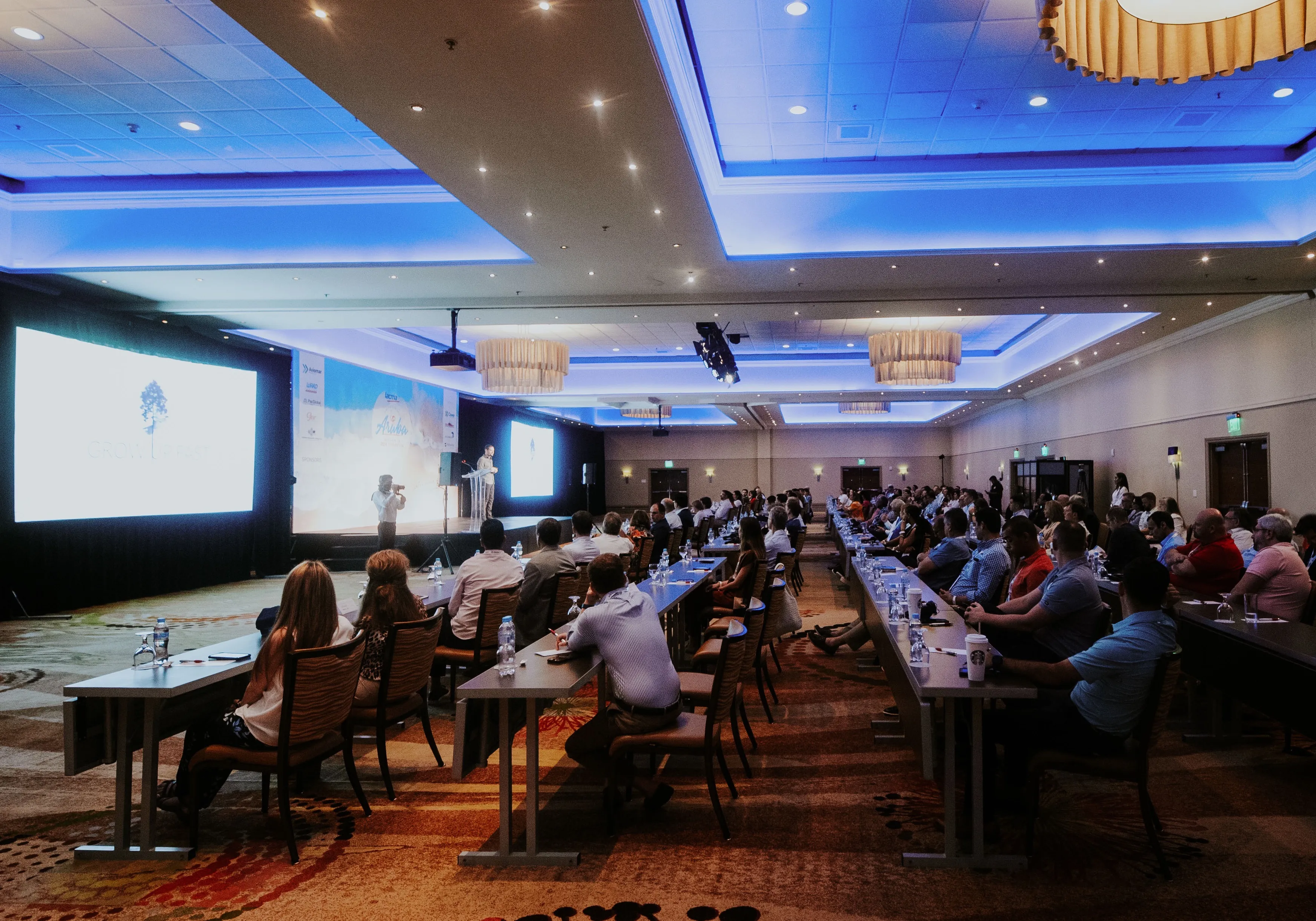 A wide shot of a professional conference with an audience seated at long tables listening to a speaker on a brightly lit stage with large presentation screens.