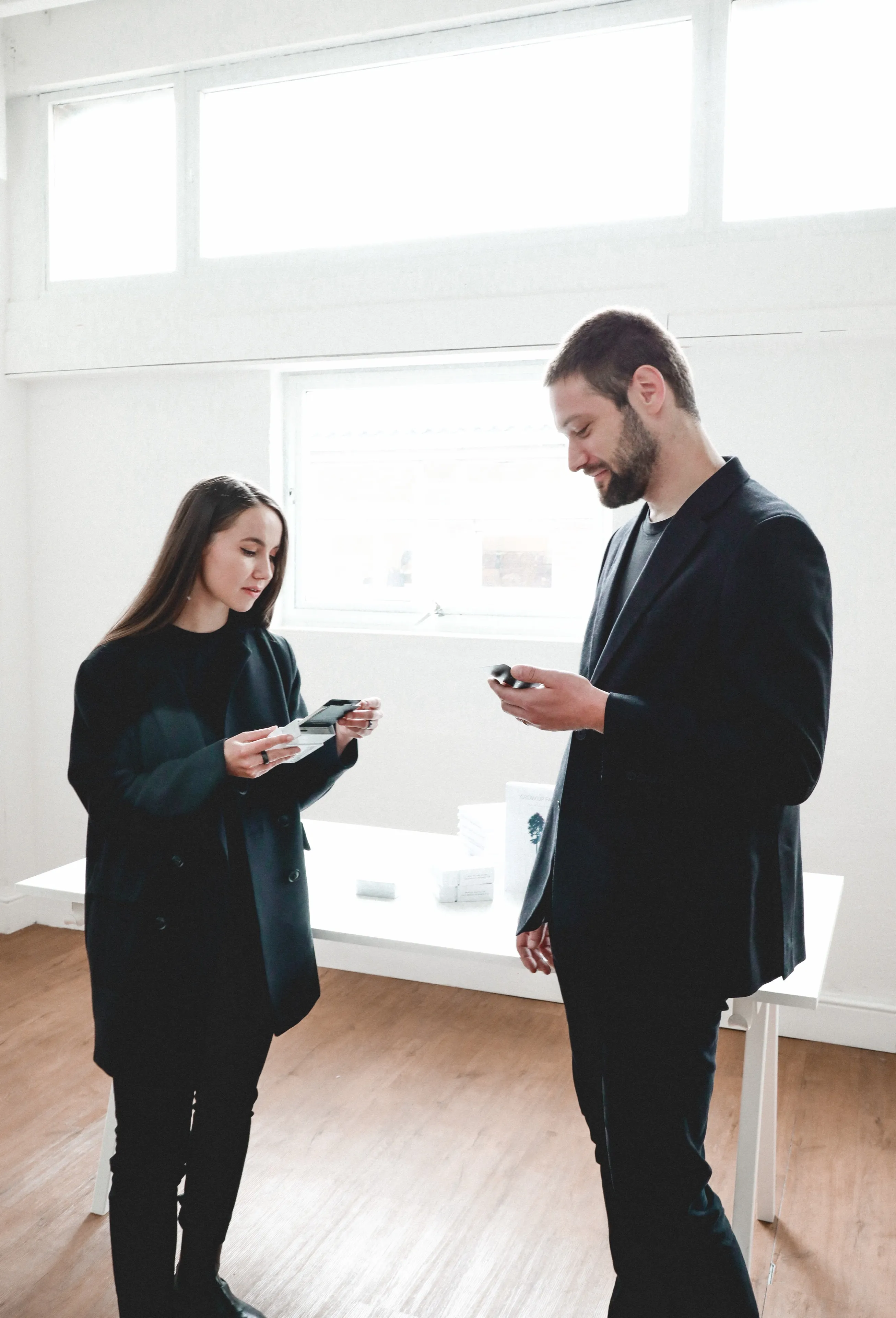 Zach and Rita wearing black blazers stand in a bright, minimalist room, both looking down at their AI Conversation Starter cards. A white table with white boxes is visible in the background.