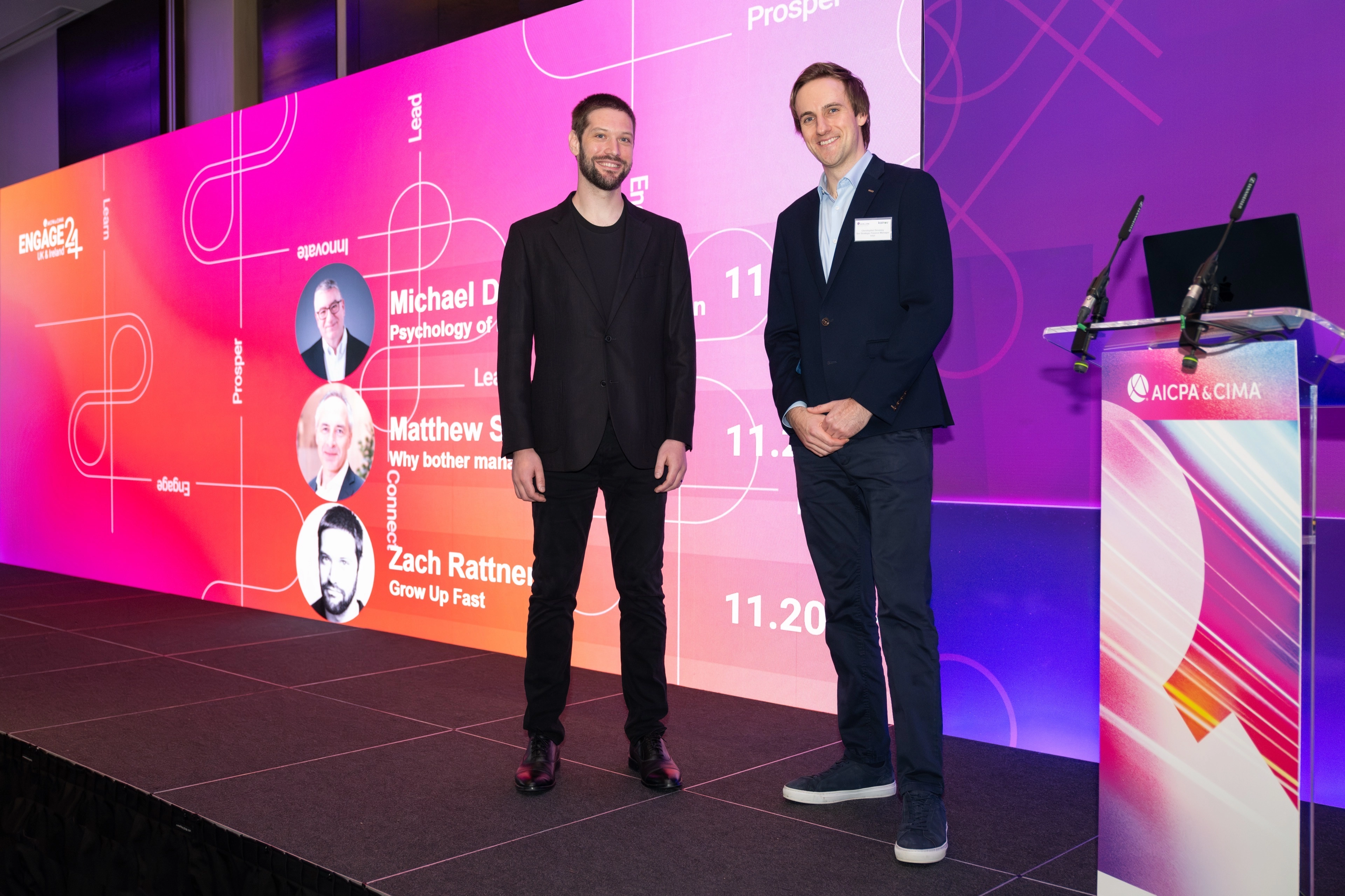 Two professional men posing on a conference stage in front of a brightly colored digital backdrop displaying speaker schedules, with a podium visible on the right.