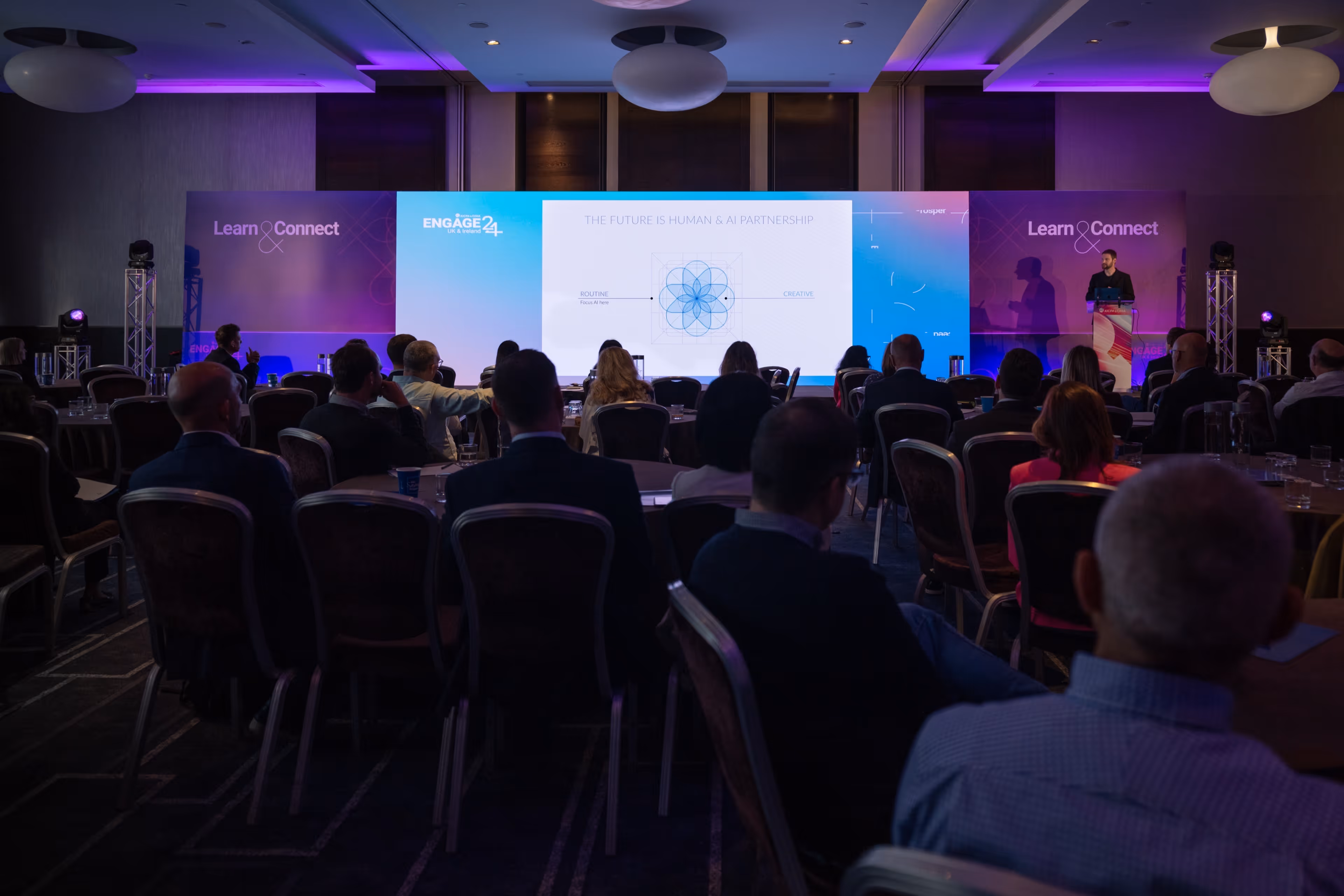 A wide shot from the back of a seated audience at a professional conference, watching a speaker on a brightly lit stage presenting a slide titled 'THE FUTURE IS HUMAN & AI PARTNERSHIP'.