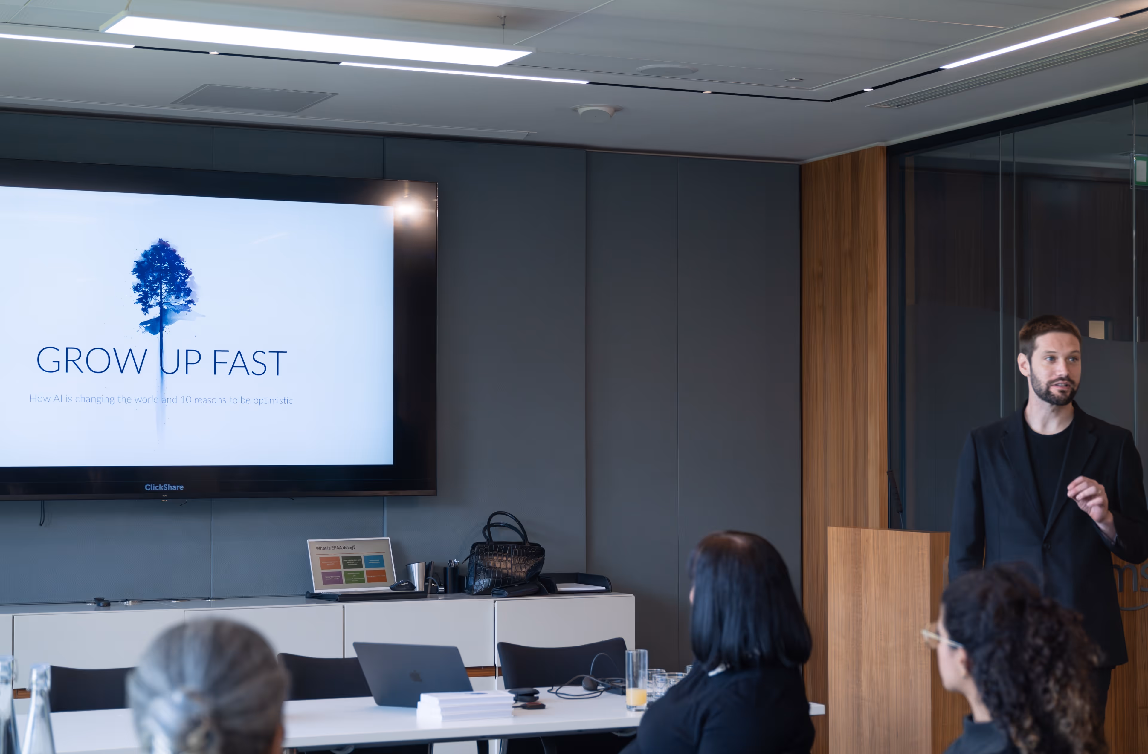 A male speaker presenting to a small seated audience in a modern conference room with a slide titled GROW UP FAST about AI visible on a large screen.