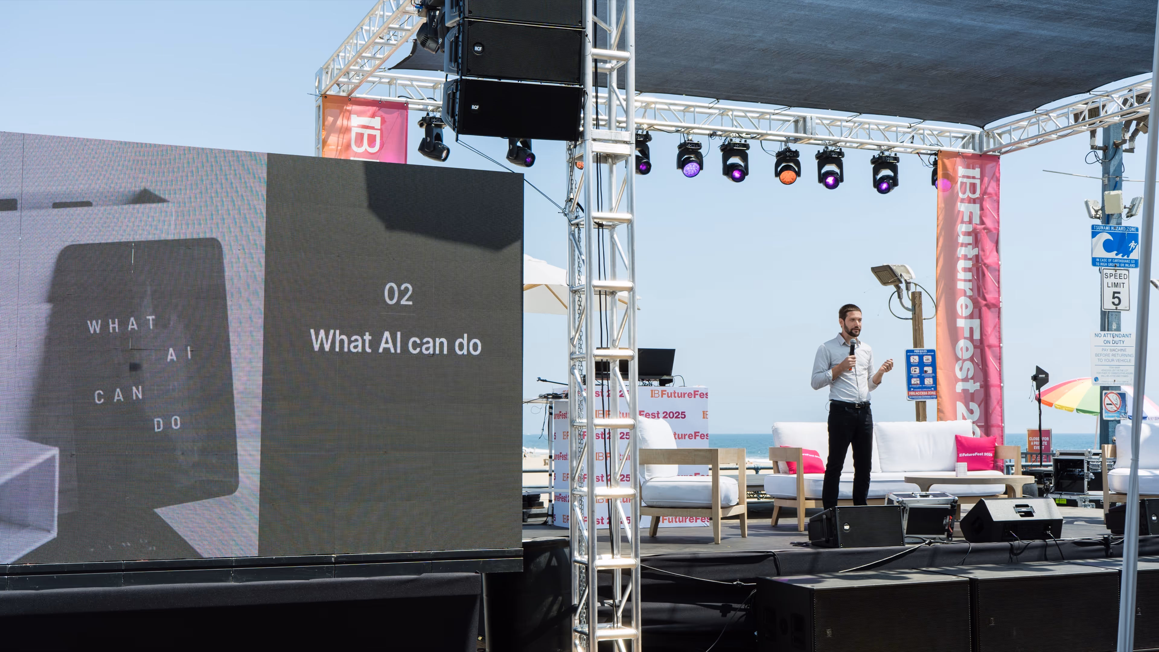 A male speaker presenting with a microphone on an outdoor stage beside a large screen displaying '02 What AI can do'.