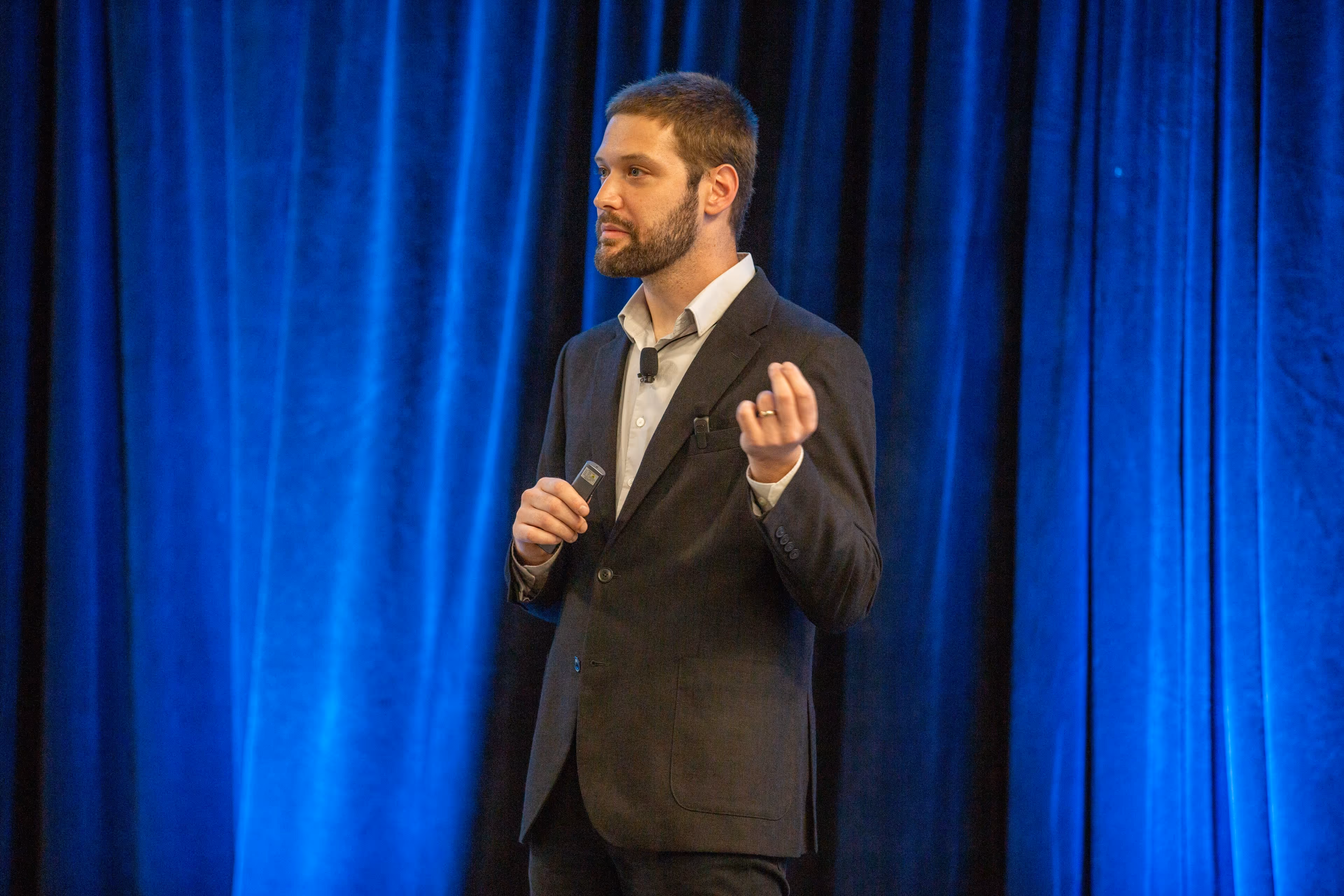 A professional man giving a presentation on stage, gesturing thoughtfully with one hand and holding a clicker in the other, set against a bright blue curtain backdrop.