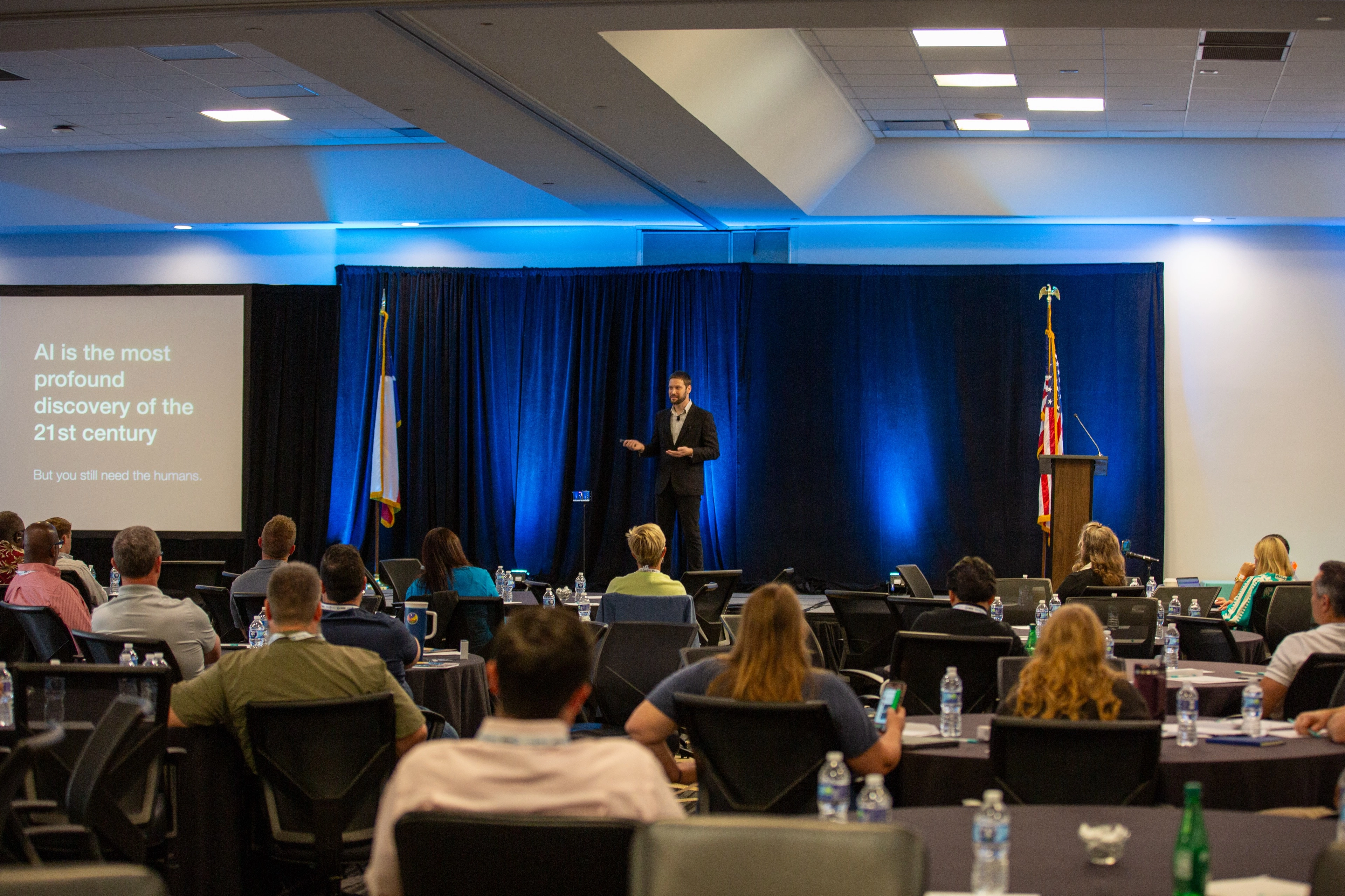 A professional presenting on stage to an audience seated at round tables, with a presentation slide about AI visible on the left.