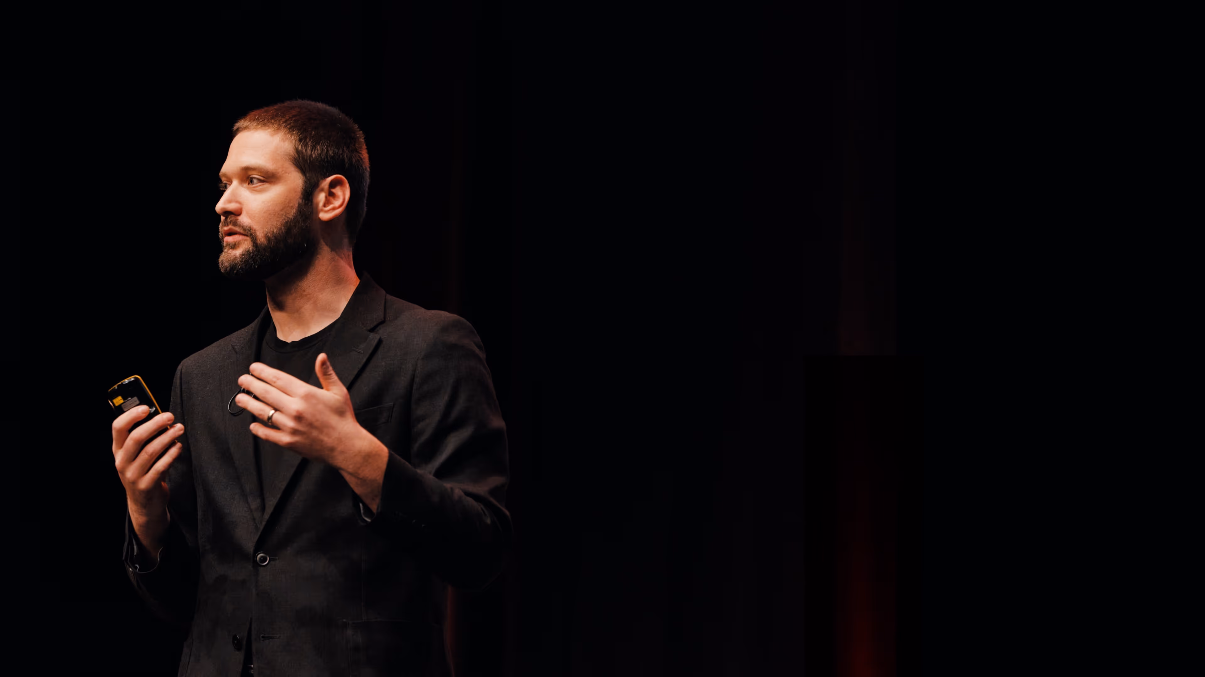 A professional man wearing a dark blazer and black t-shirt speaking on stage against a completely black background. He is holding a presentation clicker in one hand and gesturing with the other while looking off-camera.