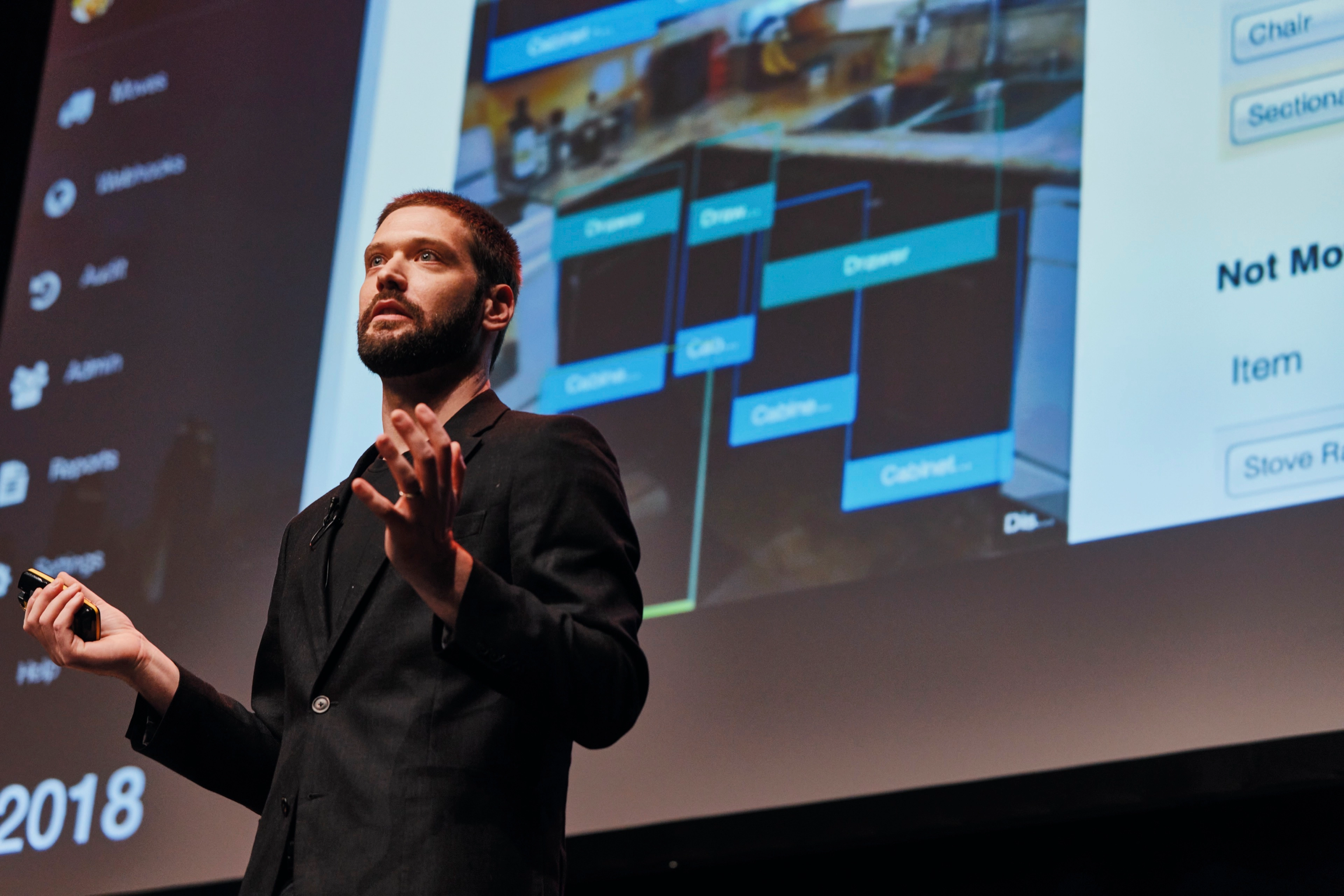 A professional man giving a presentation on stage, holding a clicker and gesturing with his hand while a technical software slide deck is projected behind him.