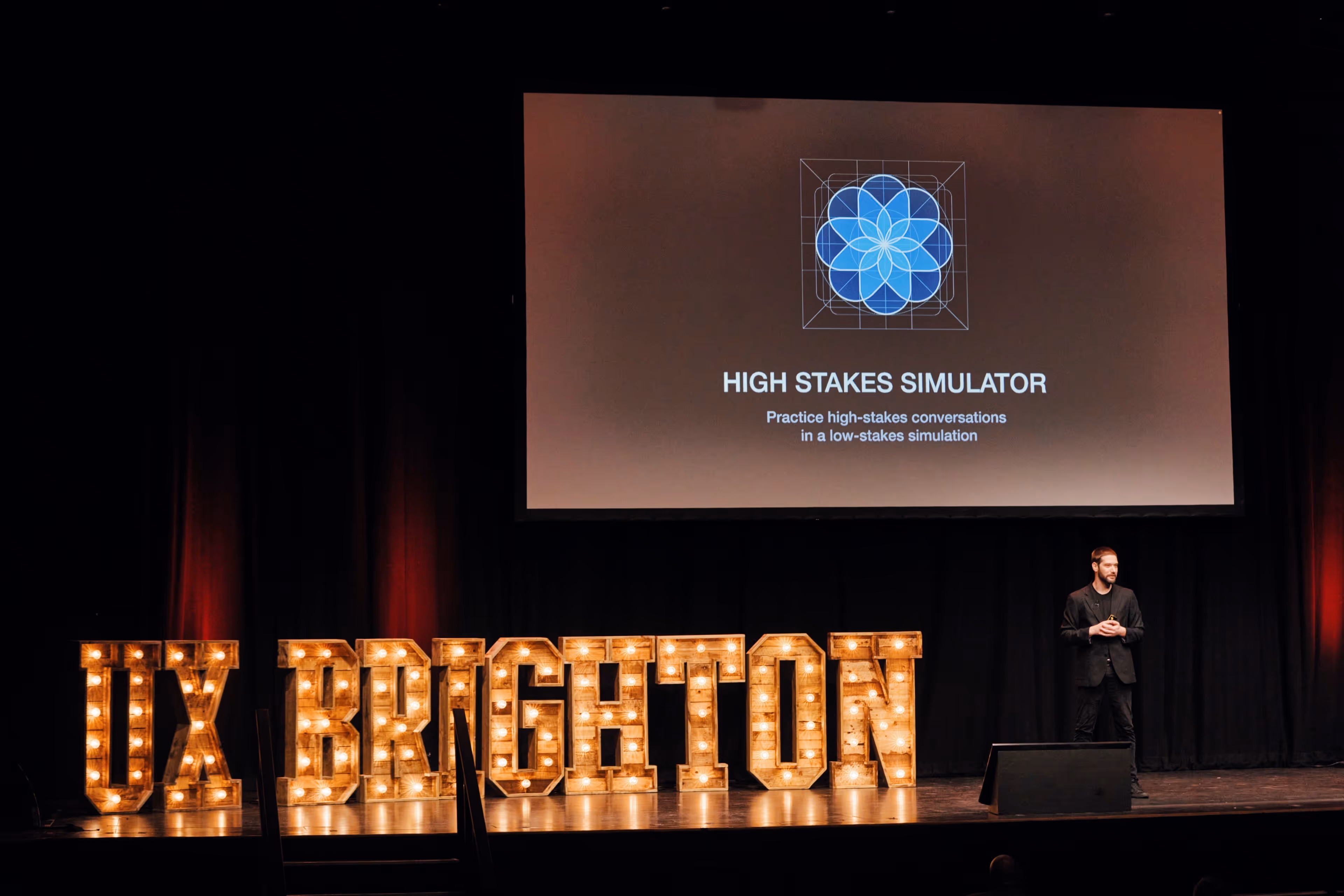 A male speaker presenting on a stage at UX Brighton. Behind him is a large screen displaying 'HIGH STAKES SIMULATOR' and a blue geometric logo. Large illuminated wooden letters spelling 'UX BRIGHTON' sit on the stage floor.