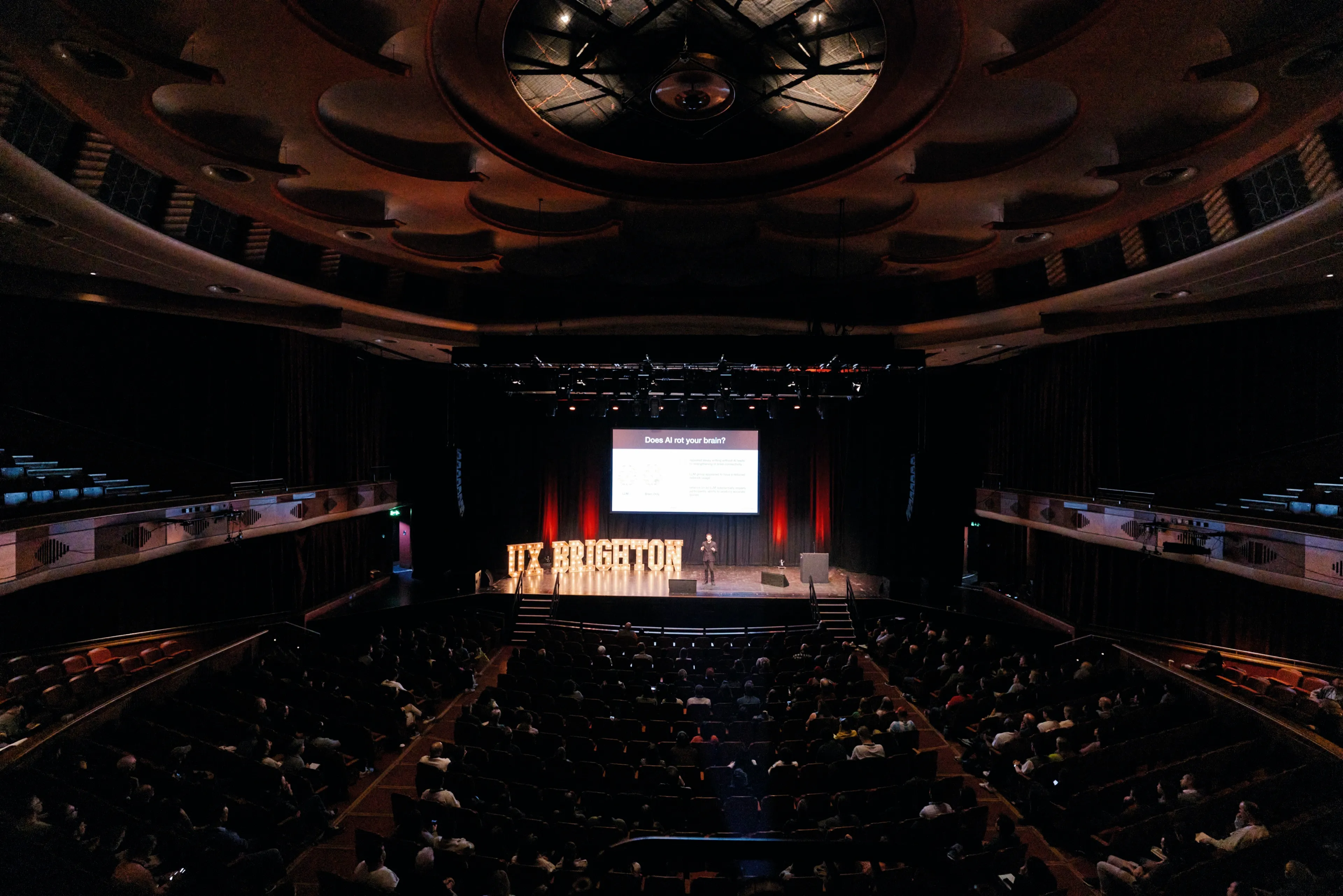 A wide shot from the back of a grand theater showing a speaker on stage presenting a slide titled 'Does AI rot your brain?' to a large audience at the UX Brighton conference.