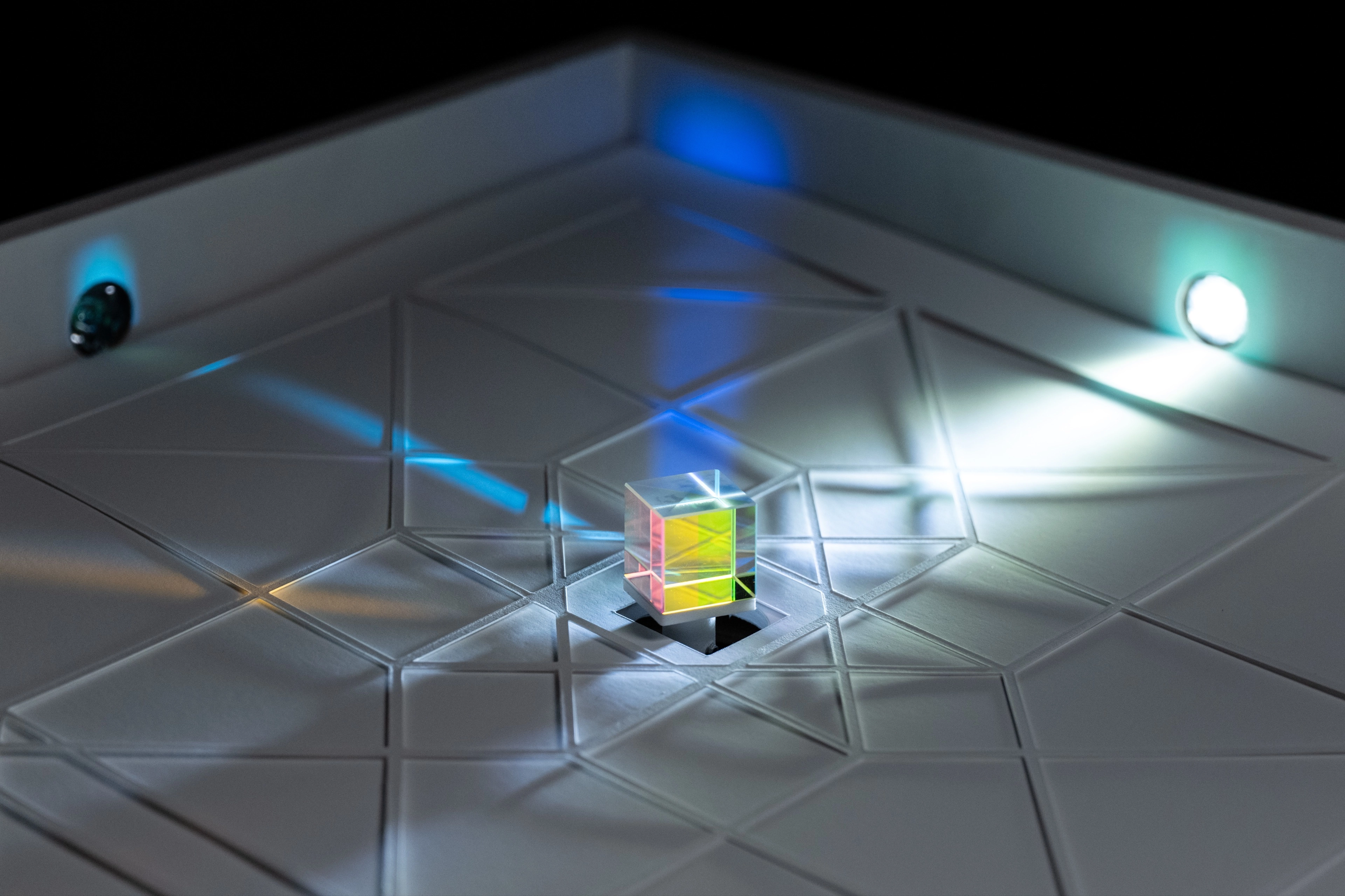 A macro shot of a dichroic prism cube on a white geometric surface, refracting a light beam into colorful reflections.