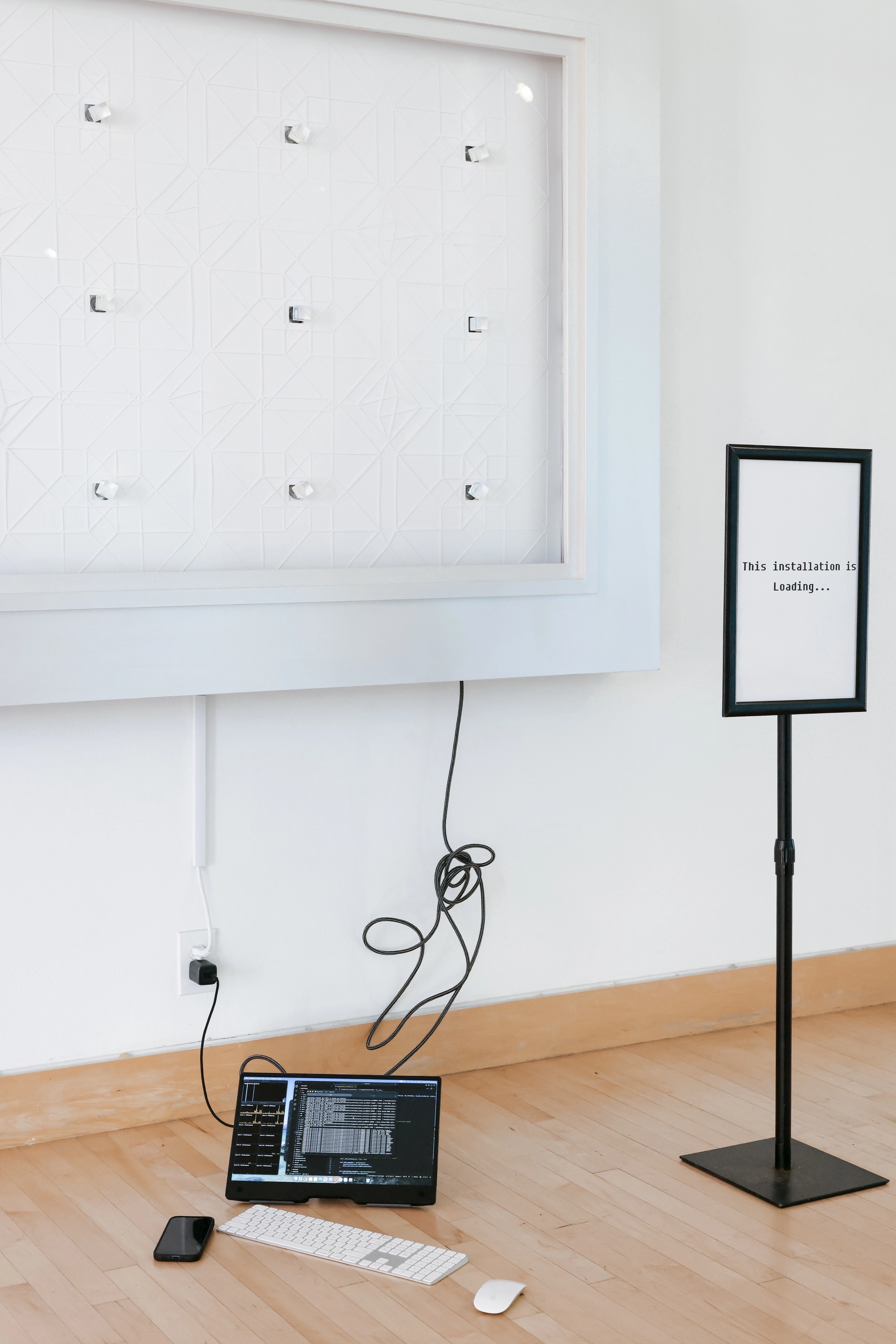 A white wall-mounted art installation connected by wires to a laptop, keyboard, and phone on the floor, next to a sign reading 'This installation is Loading...'