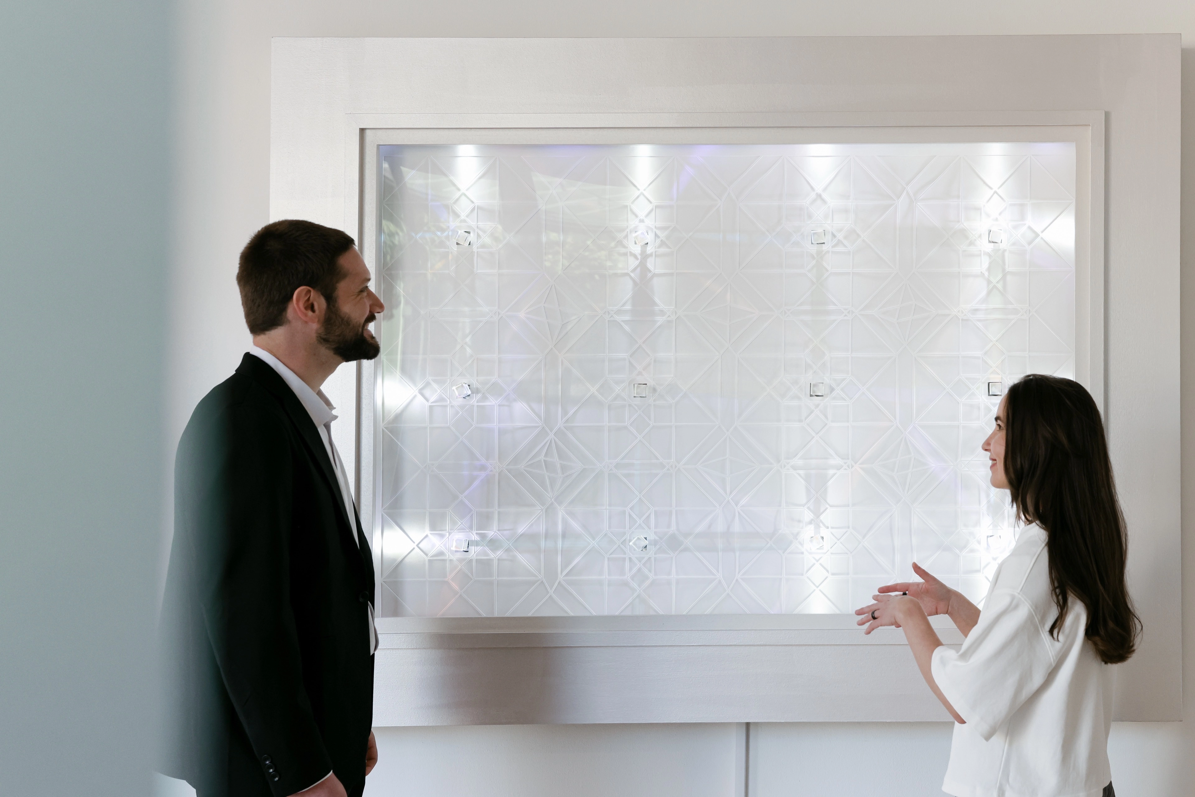 Zach in a dark suit and Rita in a white blouse are smiling and engaged in a conversation in front of a large, modern, illuminated geometric wall panel.