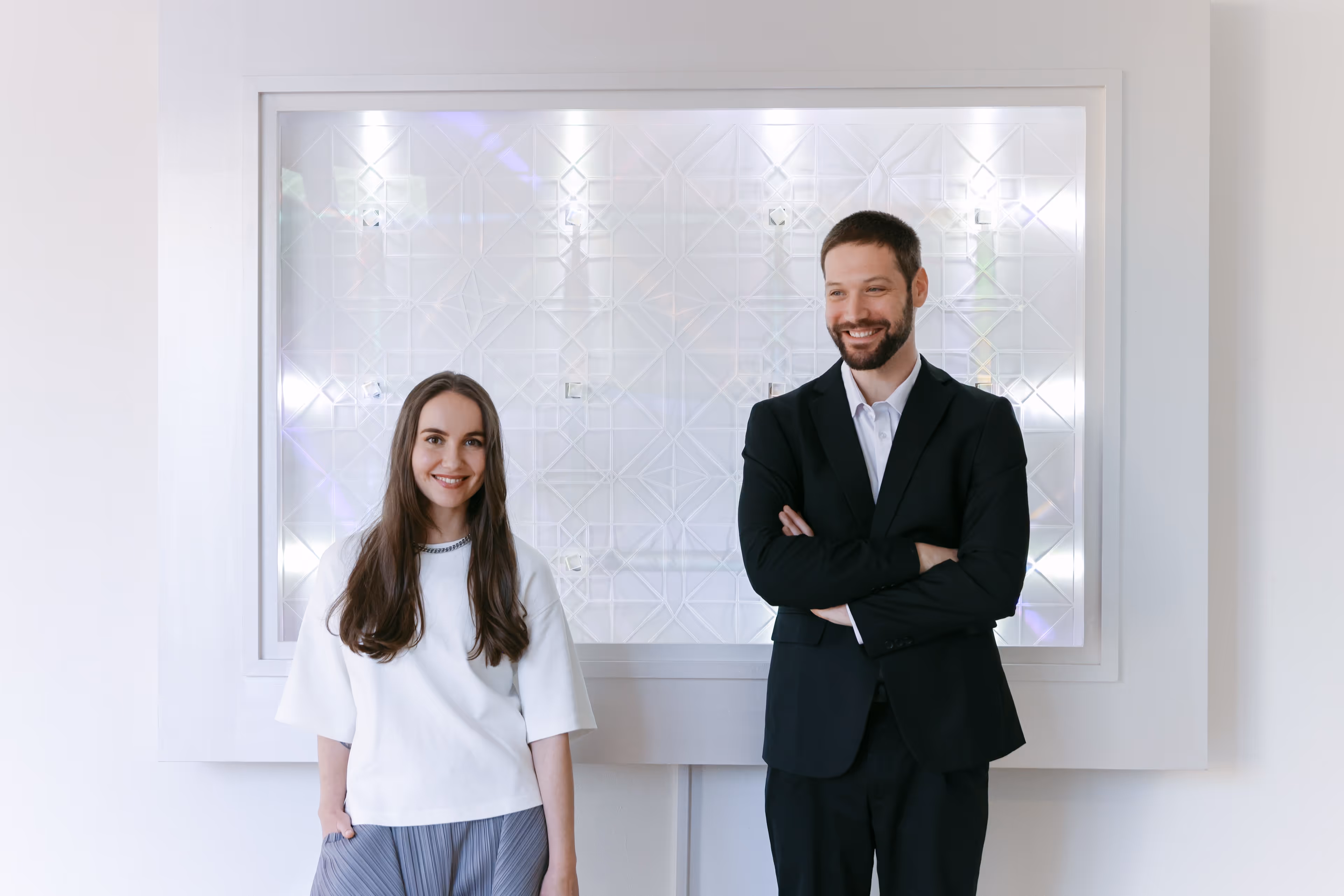 A smiling woman in a white top and a smiling man in a dark suit standing together in front of a modern, illuminated geometric wall panel.