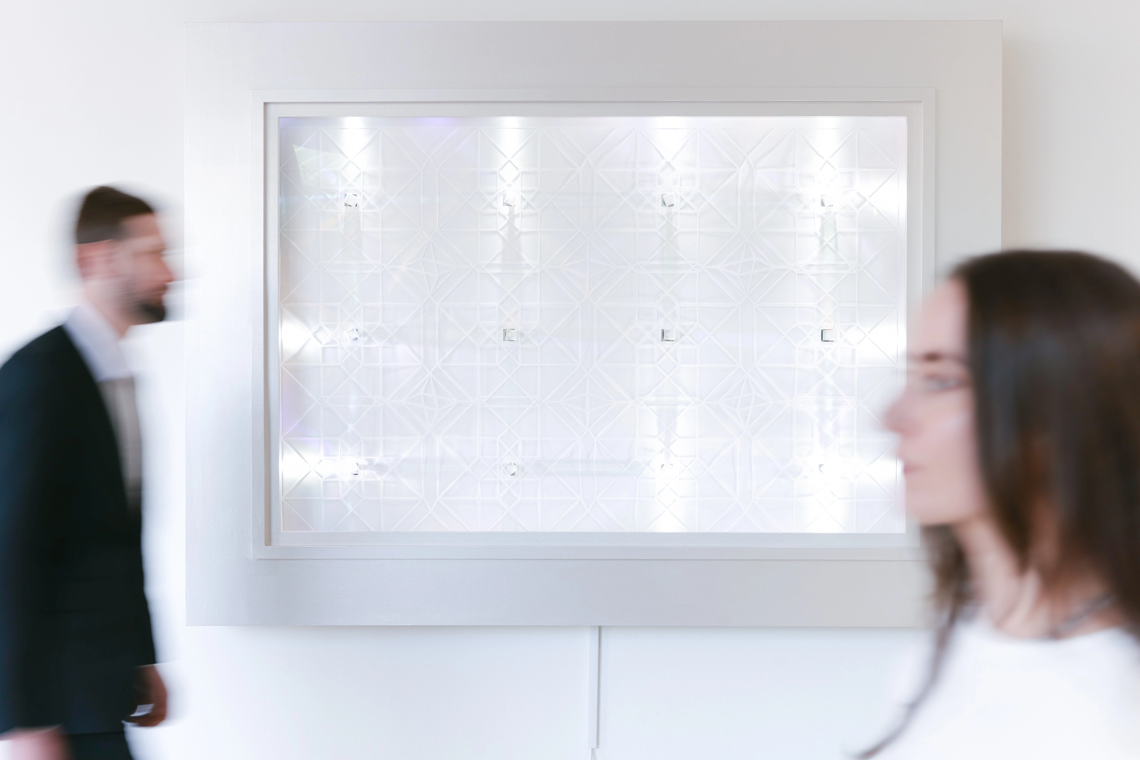 Two blurred figures in business attire walking past a large bright geometric light fixture on a minimalist white wall.